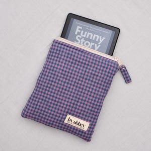 Könnte beinhalten: Eine violette und dunkel-türkisfarbene, karierte Reißverschlusstasche mit einem schwarzen E-Reader. Auf dem Bildschirm des E-Readers steht der Text Funny Story. Ein kleines rechteckiges Stoffetikett mit dem Text by abbey ist unten aufgenäht.