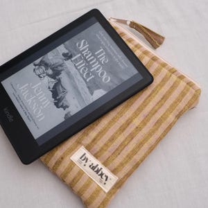 May include: A black Kindle e-reader displaying the book cover for The Shampoo Effect by Jenny Jackson, resting on a rectangular fabric pouch with a gold and light pink vertical striped pattern and a white label reading by abbey.