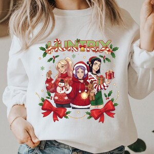 May include: White sweatshirt featuring a festive graphic with the word "HUNTRIX" in candy cane lettering. The design includes three cartoon figures in holiday attire, surrounded by holly, bows, and a string of lights. The figures are holding gifts and a reindeer.