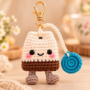 May include: A crochet keychain shaped like a tea bag with a cream-colored top, brown bottom, and two small gray feet. It features black bead eyes, pink stitched cheeks, a smiling mouth, and a bright blue circular tag attached by a cord.