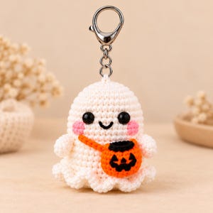 May include: A white crocheted ghost keychain featuring black bead eyes, a stitched black smile, and pink cheeks. The ghost wears a small orange crocheted pumpkin bucket with a black jack-o'-lantern face, attached to a silver metal keyring.