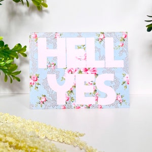 May include: A rectangular card with the words "HELL YES" in large, light pink letters. The background is a light blue floral pattern with pink roses and a gray damask design. Decorative sprigs of greenery and yellow flowers are in the background.