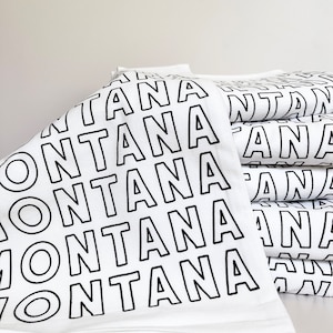 May include: A stack of white towels with the word "MONTANA" printed in bold, black capital letters. The towels are neatly folded and stacked, with the text repeated across the surface.
