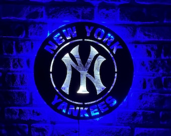 May include: A round, black sign with the words "NEW YORK YANKEES" in white lettering. The center features the team's logo in white. The sign is illuminated with blue LED lights, set against a brick wall.