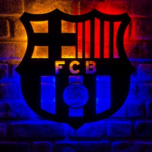 Puede incluir: Un escudo negro del FC Barcelona iluminado con luces rojas, azules y amarillas, sobre una pared de ladrillos. El escudo muestra las letras "FCB". La iluminación crea un efecto visual llamativo, ideal para la habitación de un fan.