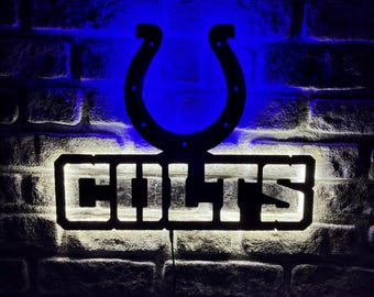 May include: A black metal Colts horseshoe and logo sign with illuminated white lettering against a brick wall. The sign is lit with blue and white lights, creating a striking contrast.