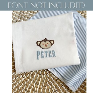 May include: White baby blanket with an embroidered monkey face and the name "PETER" in blue thread. The blanket is folded over a blue and white striped fabric, resting on a woven surface. The text "FONT NOT INCLUDED" is at the top.