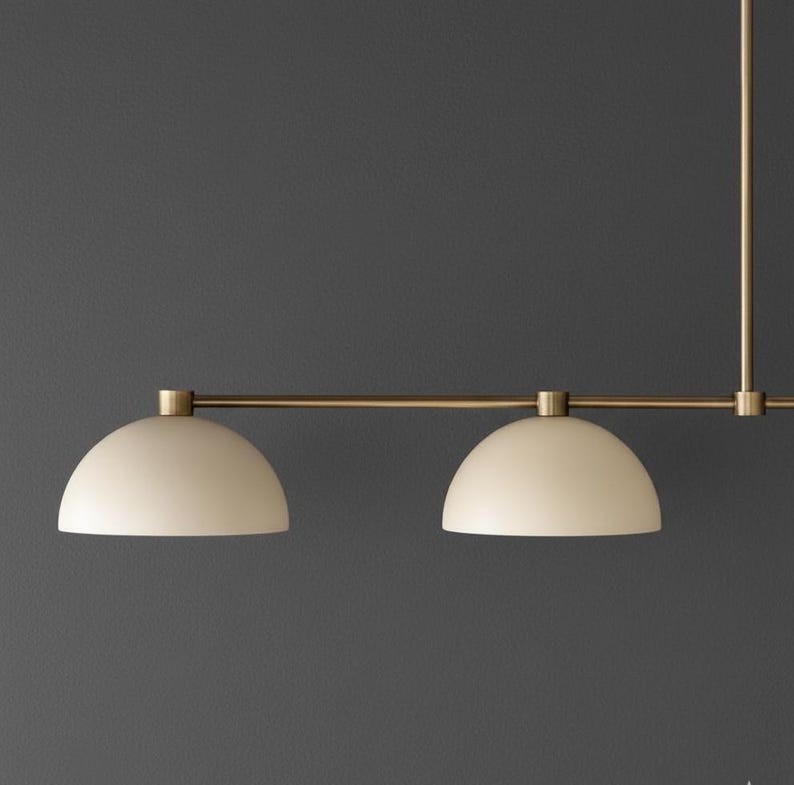 May include: A linear pendant light fixture featuring a horizontal brass rod and two downward-facing, semi-spherical off-white shades. The fixture is suspended by a vertical brass rod, showcasing a clean, symmetrical design with metallic accents.