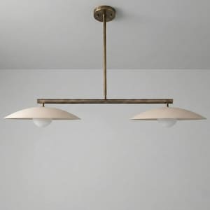 May include: A modern, two-light pendant light fixture. The fixture has a brass finish with two off-white, saucer-shaped shades. Each shade has a white, spherical light bulb. The fixture is suspended from the ceiling.