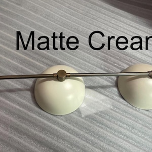 May include: A lighting fixture featuring two matte cream dome-shaped shades attached to a horizontal metallic rod. The metal components have a brushed finish, and the text "Matte Cream" is clearly visible above the two light shades.