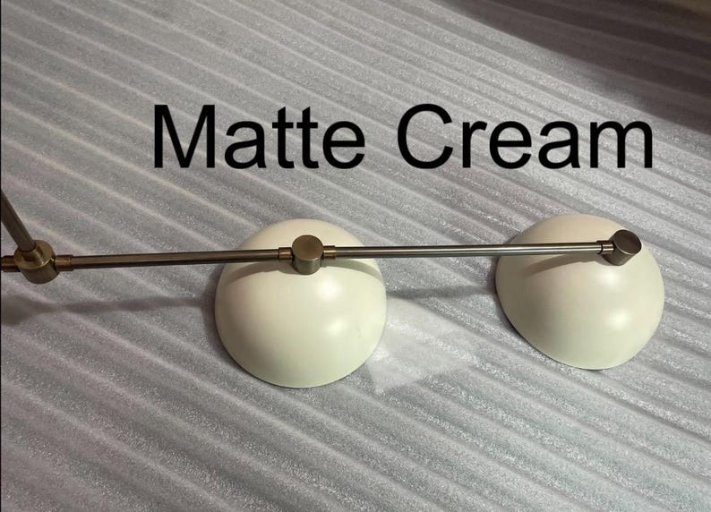 May include: A lighting fixture featuring two matte cream dome-shaped shades attached to a horizontal metallic rod. The metal components have a brushed finish, and the text "Matte Cream" is clearly visible above the two light shades.