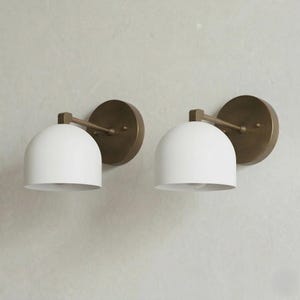 Op de afbeelding: Twee wandkandelaars met witte, koepelvormige kappen en messingkleurige montagehardware. De wandkandelaars hebben een eenvoudig, modern ontwerp, perfect voor het toevoegen van sfeerverlichting. De ronde achterplaten zijn aan de muur bevestigd.