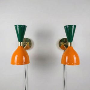 Op de afbeelding: Twee wandkandelaars in mid-century modern stijl. Elke wandkandelaar heeft een oranje, klokvormige kap, een groene, kegelvormige bovenkant en een messingkleurige montageplaat. De wandkandelaars zijn verbonden met een zwart netsnoer.