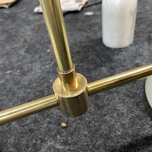 May include: A brass-colored metal light fixture component featuring a central cylindrical connector joining three thin, straight rods. The finish is smooth and reflective, with a polished metallic appearance throughout the structural assembly.