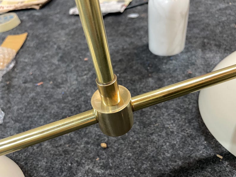 May include: A brass-colored metal light fixture component featuring a central cylindrical connector joining three thin, straight rods. The finish is smooth and reflective, with a polished metallic appearance throughout the structural assembly.