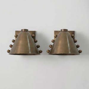 Puede incluir: Dos apliques de pared de color bronce. Cada aplique tiene una pantalla cónica con una serie de pequeños elementos decorativos redondos. Los apliques están montados sobre placas cuadradas. El diseño es moderno y elegante.