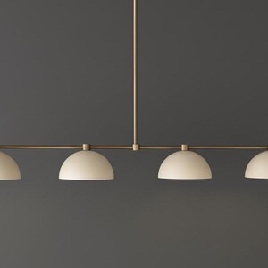 May include: A linear pendant light fixture featuring a thin, horizontal brass-toned metal rod suspended from a central vertical stem. Four matte off-white dome-shaped shades are evenly spaced along the rod, each pointing downward.