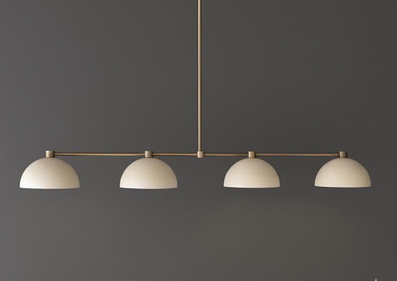 May include: A linear pendant light fixture featuring a thin, horizontal brass-toned metal rod suspended from a central vertical stem. Four matte off-white dome-shaped shades are evenly spaced along the rod, each pointing downward.