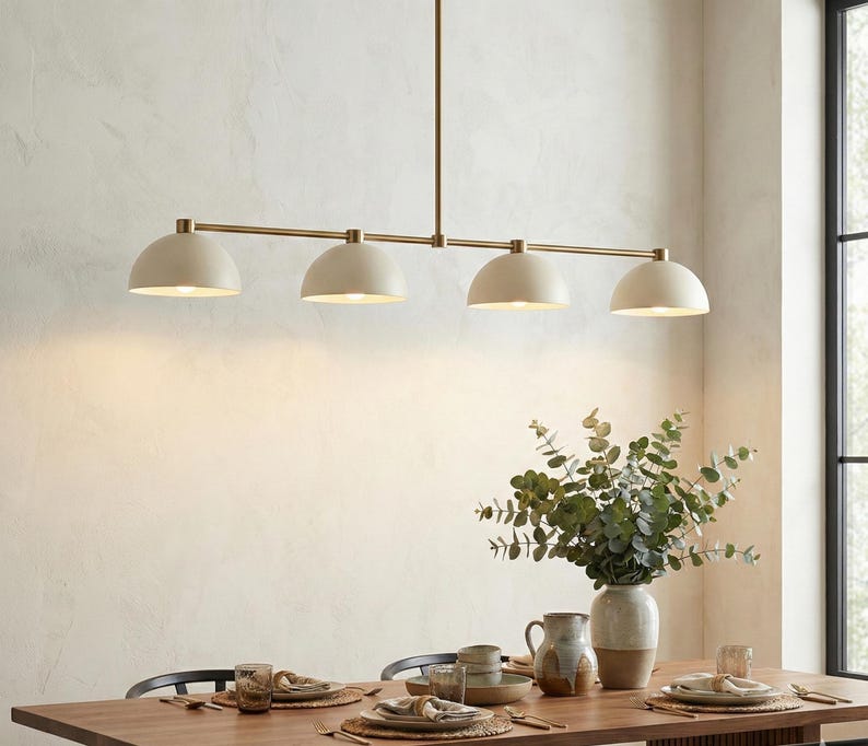 May include: A linear pendant light fixture featuring four matte cream-colored dome shades suspended from a thin, horizontal brass-toned metal rod. The fixture is mounted to the ceiling by a single vertical rod, providing downward illumination.