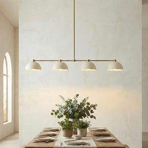 May include: A linear pendant light fixture featuring a long horizontal brass rod suspended from the ceiling, holding four matte off-white dome-shaped shades that direct light downward over a wooden dining table set with plates and greenery.