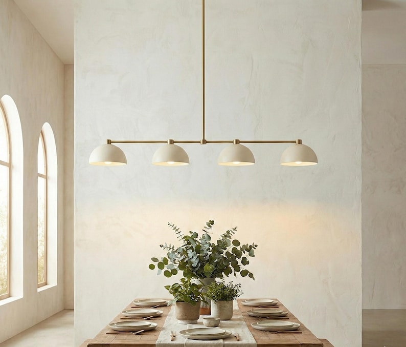 May include: A linear pendant light fixture featuring a long horizontal brass rod suspended from the ceiling, holding four matte off-white dome-shaped shades that direct light downward over a wooden dining table set with plates and greenery.