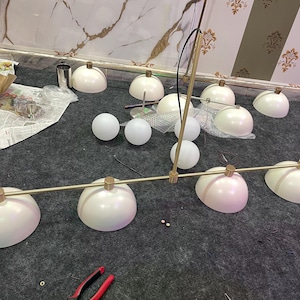 May include: A linear chandelier featuring a long, thin, brushed gold metal rod with four matte white dome-shaped shades attached at intervals. The fixture includes several additional loose white dome shades and spherical glass globes.