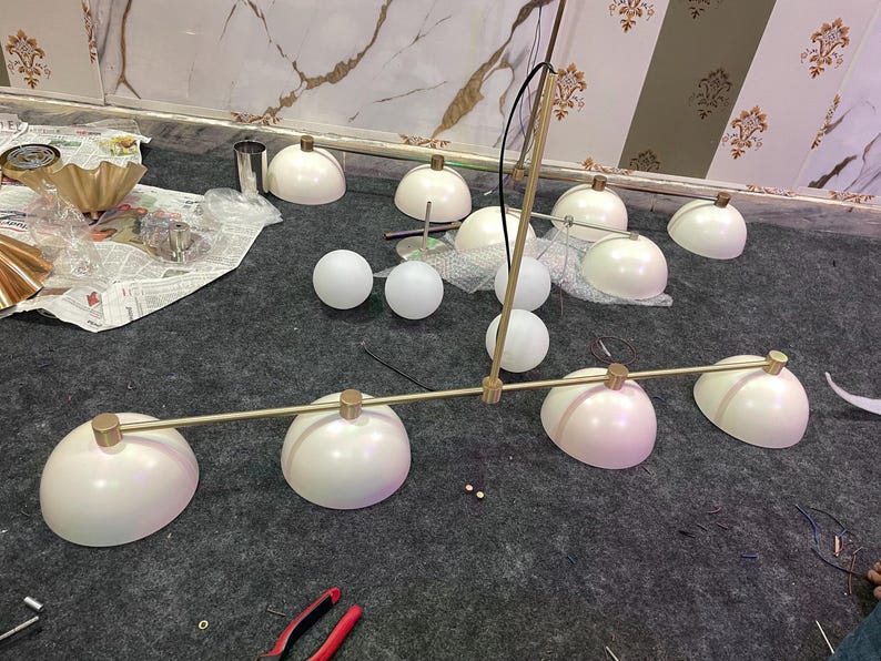 May include: A linear chandelier featuring a long, thin, brushed gold metal rod with four matte white dome-shaped shades attached at intervals. The fixture includes several additional loose white dome shades and spherical glass globes.