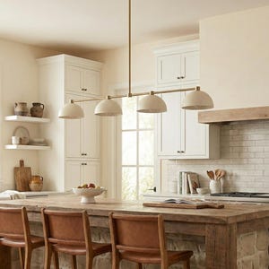 May include: A linear pendant light fixture featuring a long, thin brass rod suspended from the ceiling, holding four evenly spaced, matte off-white dome-shaped shades that direct light downward over a wooden kitchen island with leather chairs.