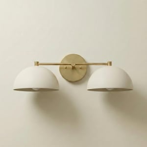 May include: A wall-mounted light fixture featuring a horizontal brass bar connected to a circular brass wall plate. Two matte off-white dome-shaped shades are attached to the ends of the bar, pointing downward to provide focused illumination.
