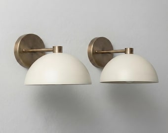 May include: Two wall sconces with a cream-colored, dome-shaped shade and a brass-colored mounting plate and arm. The sconces are mounted on a light gray wall, providing a warm, inviting glow. These fixtures are ideal for adding ambient lighting to any room.