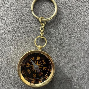May include: A gold-coloured compass keyring with a black face and white and gold markings. The compass has a ring for attaching keys and a small chain. The compass face displays directional markings.