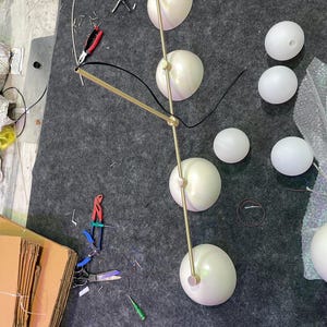 May include: A multi-light chandelier featuring a long, thin, gold-toned metal rod with four attached hemispherical light shades. The fixture includes several additional detached white spherical glass globes and black electrical wiring components.