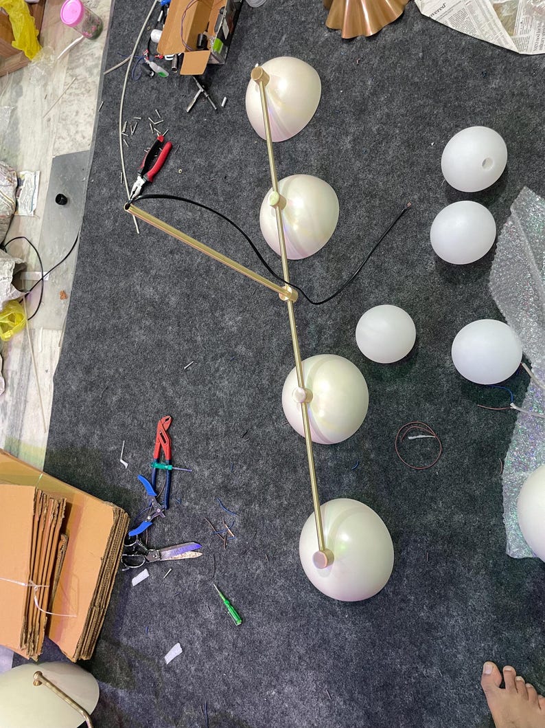 May include: A multi-light chandelier featuring a long, thin, gold-toned metal rod with four attached hemispherical light shades. The fixture includes several additional detached white spherical glass globes and black electrical wiring components.