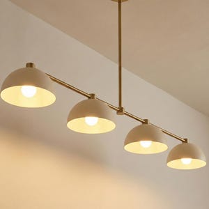 May include: A linear chandelier featuring four matte beige dome-shaped shades attached to a central brass-toned metal rod. Each shade houses a single glowing light bulb, creating a downward-facing illumination pattern from the horizontal fixture.