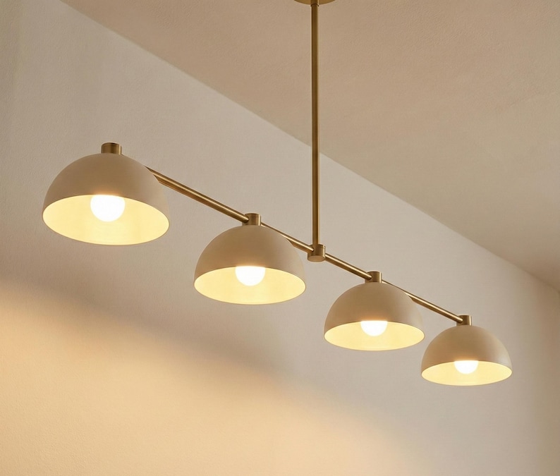 May include: A linear chandelier featuring four matte beige dome-shaped shades attached to a central brass-toned metal rod. Each shade houses a single glowing light bulb, creating a downward-facing illumination pattern from the horizontal fixture.