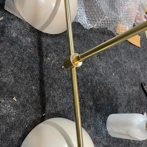 May include: A multi-arm light fixture featuring two matte cream-colored dome shades and a central brass-toned metal frame. The structure includes adjustable joints and a cylindrical white marble-patterned accent piece attached to one of the arms.