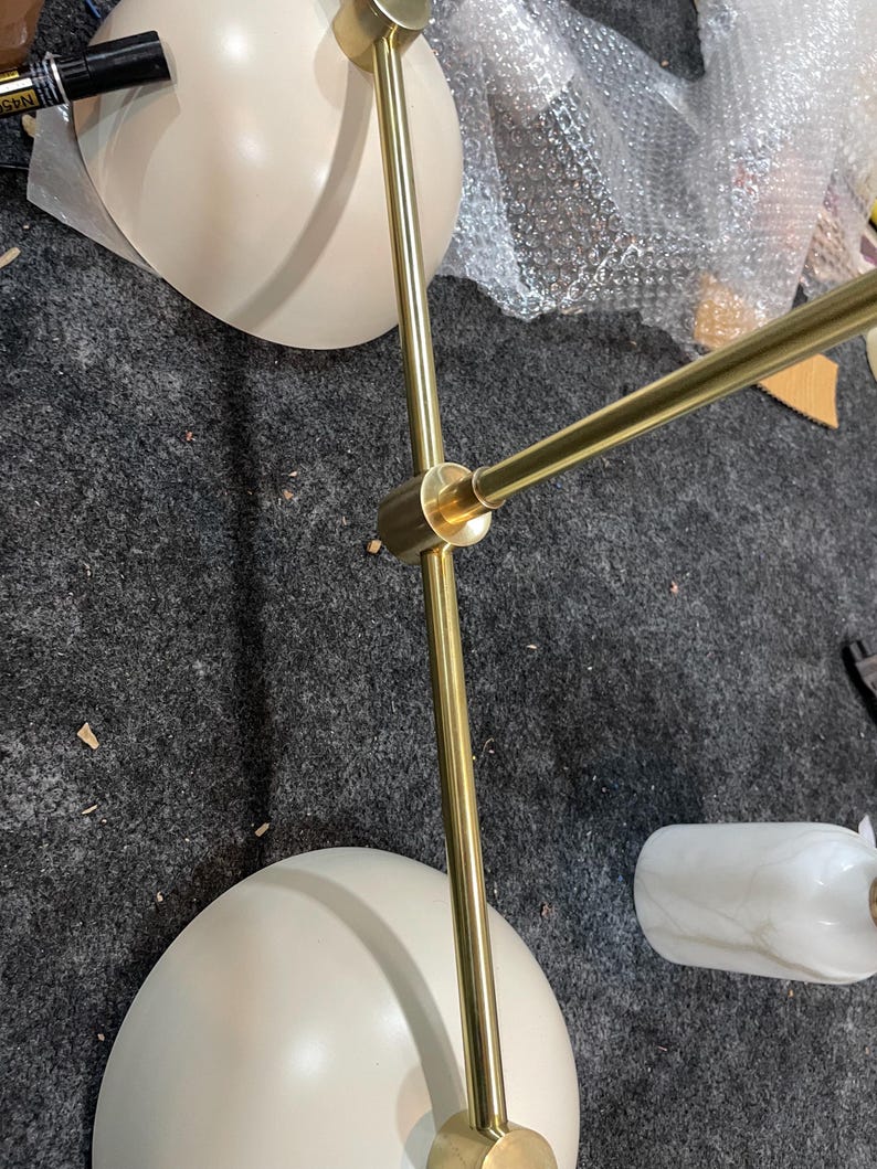 May include: A multi-arm light fixture featuring two matte cream-colored dome shades and a central brass-toned metal frame. The structure includes adjustable joints and a cylindrical white marble-patterned accent piece attached to one of the arms.