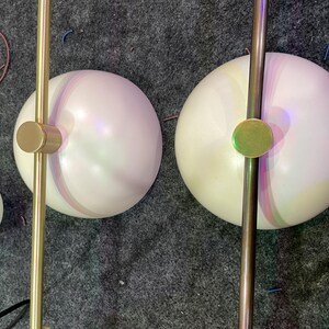 May include: Two spherical white glass light shades attached to a straight gold-toned metal rod. Each shade features a central circular gold-toned metal cap on top. A third smaller, detached white glass sphere is positioned to the left side.