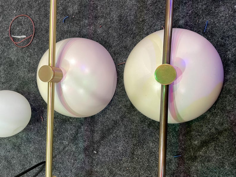 May include: Two spherical white glass light shades attached to a straight gold-toned metal rod. Each shade features a central circular gold-toned metal cap on top. A third smaller, detached white glass sphere is positioned to the left side.