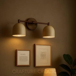 May include: A two-light wall sconce featuring a dark bronze horizontal bar and circular wall mount. Two matte beige dome-shaped shades hang downward, emitting a warm glow. Below the fixture are two framed prints with light wood frames.