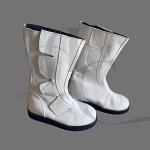 Könnte beinhalten: Ein Paar weiße Lederstiefel mit mittelhohem Schaft. Die Stiefel haben ein einzigartiges Design mit eckigen Aussparungen und einer schwarzen Sohle. Die Stiefel sind vor grauem Hintergrund abgebildet.