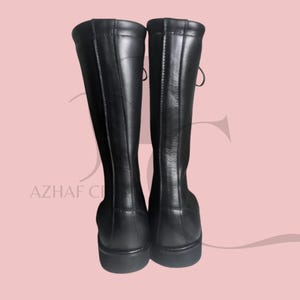 K&ouml;nnte beinhalten: Ein Paar schwarze kniehohe Stiefel. Die Stiefel bestehen aus einem glatten, gl&auml;nzenden Material und haben eine runde Spitze. Die Stiefel haben ein Schn&uuml;rdesign auf der R&uuml;ckseite und einen kleinen Absatz. Die Stiefel werden vor einem hellrosa Hintergrund pr&auml;sentiert.