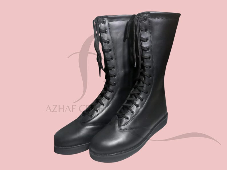 K&ouml;nnte beinhalten: Ein Paar schwarze, kniehohe Stiefel mit Schn&uuml;rdesign. Die Stiefel haben ein glattes Leder-Aussehen und eine flache Sohle. Das Bild enth&auml;lt den Text "AZHAF CREATIONS" in der unteren linken Ecke.