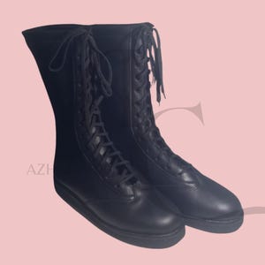 K&ouml;nnte beinhalten: Ein Paar schwarze, kniehohe Stiefel mit Schn&uuml;rdesign. Die Stiefel haben ein glattes, lederartiges Aussehen und eine flache Sohle. Die Stiefel werden vor einem hellrosa Hintergrund pr&auml;sentiert.