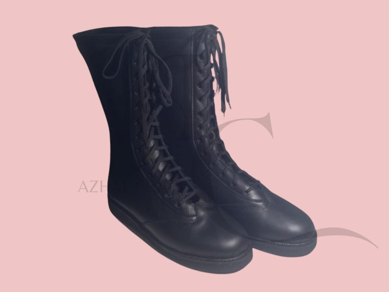 K&ouml;nnte beinhalten: Ein Paar schwarze, kniehohe Stiefel mit Schn&uuml;rdesign. Die Stiefel haben ein glattes, lederartiges Aussehen und eine flache Sohle. Die Stiefel werden vor einem hellrosa Hintergrund pr&auml;sentiert.