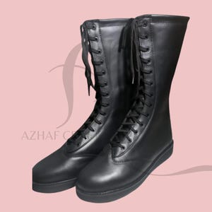 K&ouml;nnte beinhalten: Ein Paar schwarze, kniehohe Stiefel mit Schn&uuml;rdesign. Die Stiefel haben ein glattes Leder-Aussehen und eine flache Sohle. Das Bild ist vor einem hellrosa Hintergrund platziert. Der Markenname "AZHAF CREATIONS" ist sichtbar.