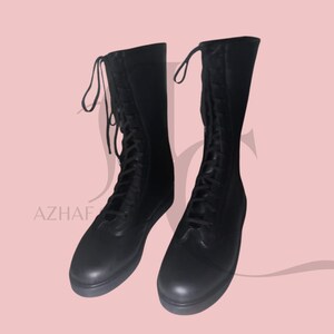 K&ouml;nnte beinhalten: Ein Paar schwarze, kniehohe Stiefel mit Schn&uuml;rdesign. Die Stiefel haben eine runde Spitze und eine glatte, matte Oberfl&auml;che. Die Schn&uuml;rsenkel reichen bis zum oberen Rand des Stiefels. Die Stiefel werden vor einem hellrosa Hintergrund pr&auml;sentiert.