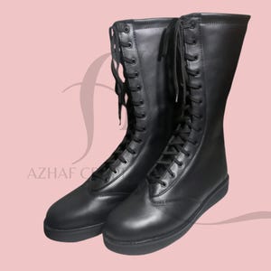 K&ouml;nnte beinhalten: Ein Paar schwarze, kniehohe Stiefel mit Schn&uuml;rdesign. Die Stiefel sind aus glattem Material und haben eine flache Sohle. Die Stiefel werden vor einem rosa Hintergrund pr&auml;sentiert. Der Text "AZHAF CREATIONS" ist sichtbar.