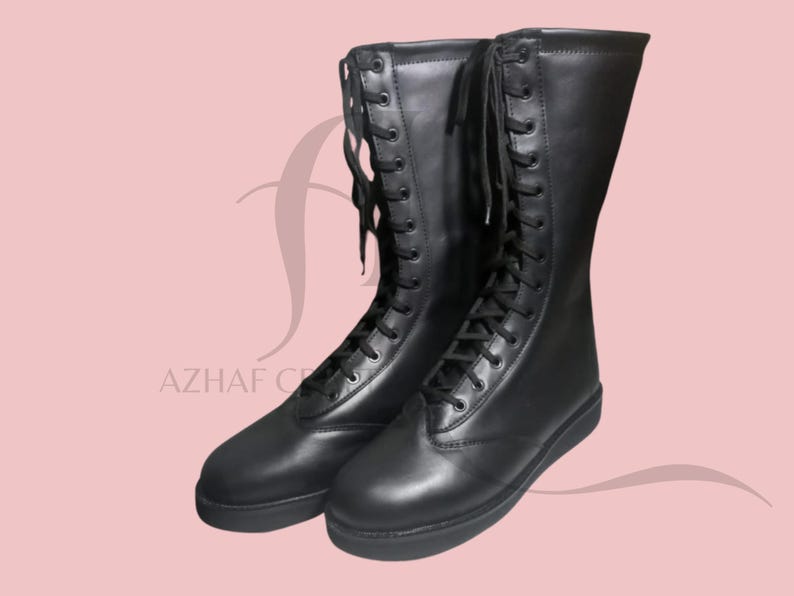 K&ouml;nnte beinhalten: Ein Paar schwarze, kniehohe Stiefel mit Schn&uuml;rdesign. Die Stiefel sind aus glattem Material und haben eine flache Sohle. Die Stiefel werden vor einem rosa Hintergrund pr&auml;sentiert. Der Text "AZHAF CREATIONS" ist sichtbar.