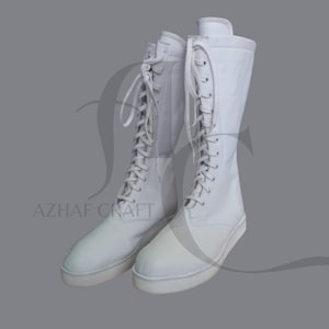 Könnte beinhalten: Ein Paar weiße, kniehohe Stiefel mit Schnürdesign. Die Stiefel haben eine flache Sohle und eine runde Spitze. Der Text "AZHAF CRAFT" ist auf der Seite der Stiefel zu sehen.